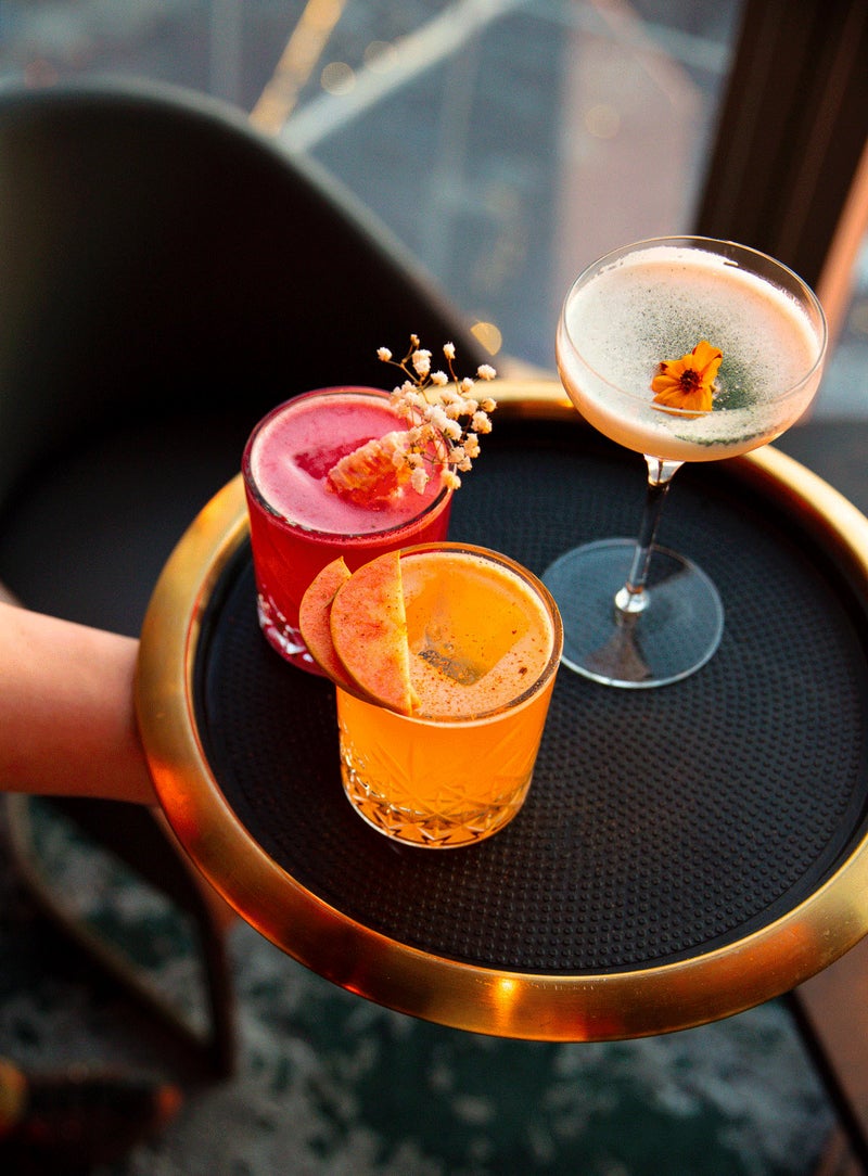 Three colorful cocktails on a round serving tray held beside a lounge seating area.